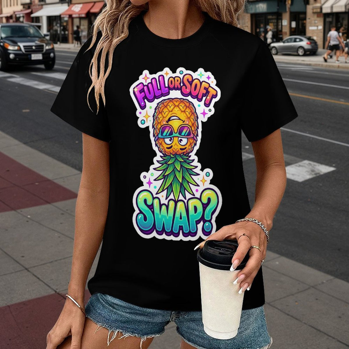 A woman wearing a black graphic t-shirt with a colorful pineapple wearing sunglasses and the text 'FULL OR SOFT SWAP?' printed on the front, standing on a city street.