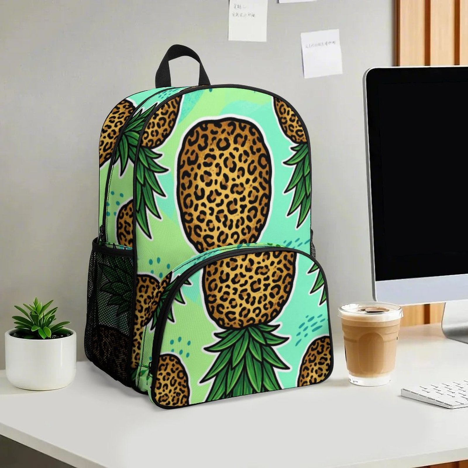 A backpack with a pineapple and leopard print pattern sits on a white desk next to a computer monitor, a small potted plant, and a cup of iced coffee.