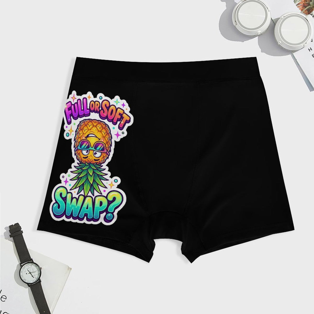 Black boxer briefs with a colorful graphic of a pineapple wearing sunglasses and a green leafy collar, with the text 'FULL OR SOFT SWAP?' in pink, purple, and blue. The underwear is displayed in a flat-lay arrangement on a white background with a watch, jars, and a card as props.