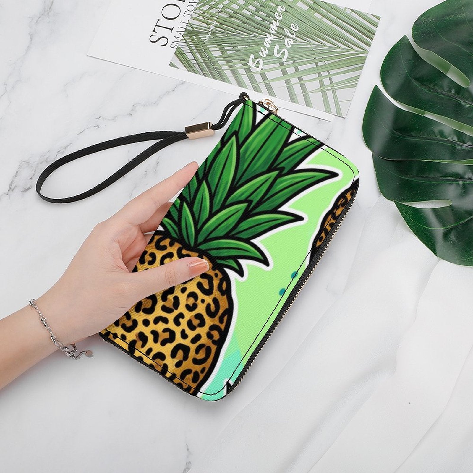 A hand holds a zip-around wallet featuring a pineapple design with a green leafy top and a leopard-print base, set on a white marble surface with decorative props.