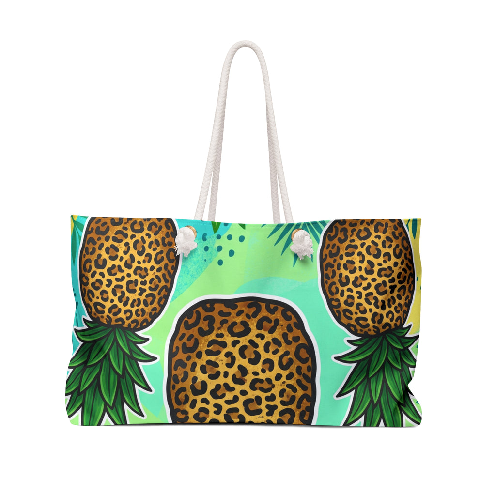 A tote bag featuring a bold graphic print of three pineapples with leopard-print bodies and green leafy tops on a light turquoise background, with white rope handles.