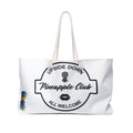 A white weekender bag with a text 'Upside Down Pineapple Club All Welcome' and a colorful pineapple illustration on it, featuring large handles.