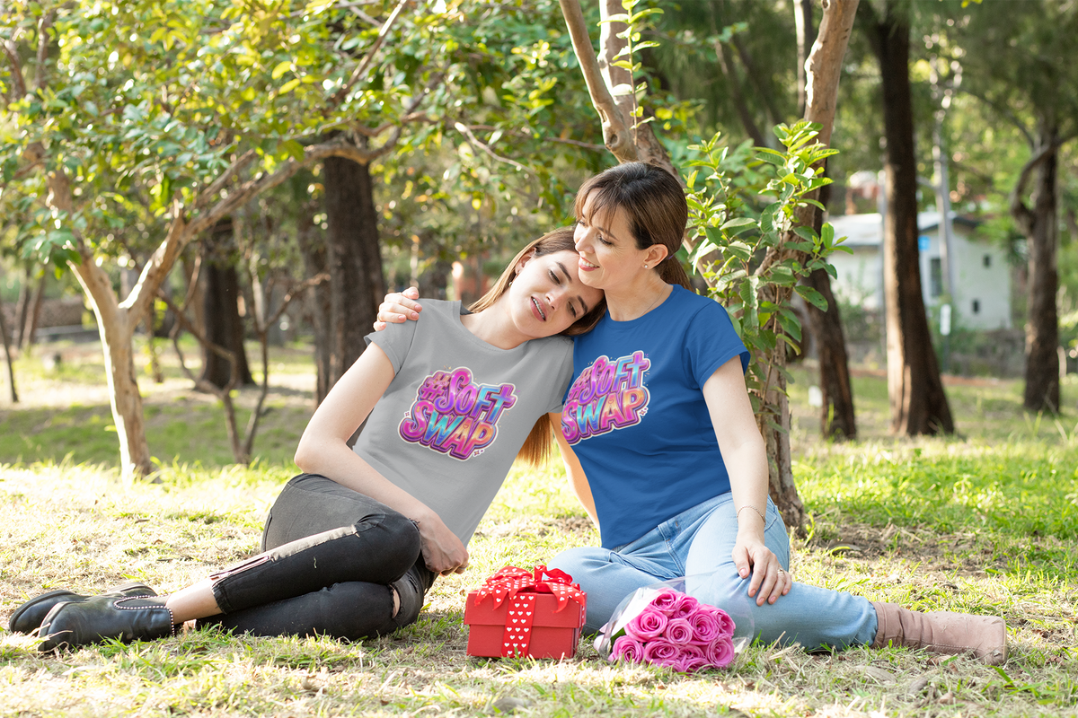 Two women sitting down on the grass in a park next to trees wearing #SoftSwap T-shirts on wearing black and blue jeans.