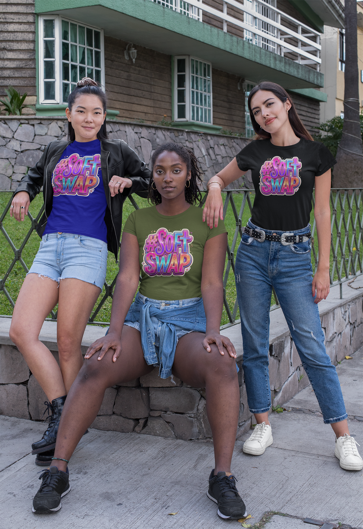 Three women posing outdoors in matching graphic t-shirts that read 'SOFTLY SWAP' in pink, purple, and blue lettering. The shirts are shown in three different colors: blue, olive green, and black.