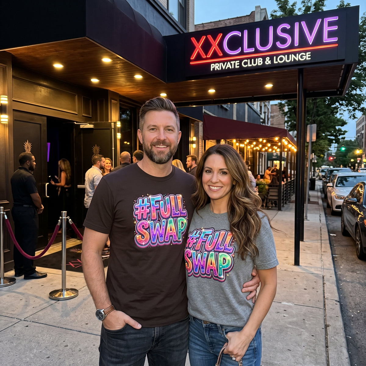 A man and a woman standing outside a nightclub or lounge entrance at dusk, both wearing matching graphic T-shirts with the text '#FULL SWAP' in a pink, blue, and yellow gradient. The man wears a dark brown T-shirt, and the woman wears a heather gray T-shirt.