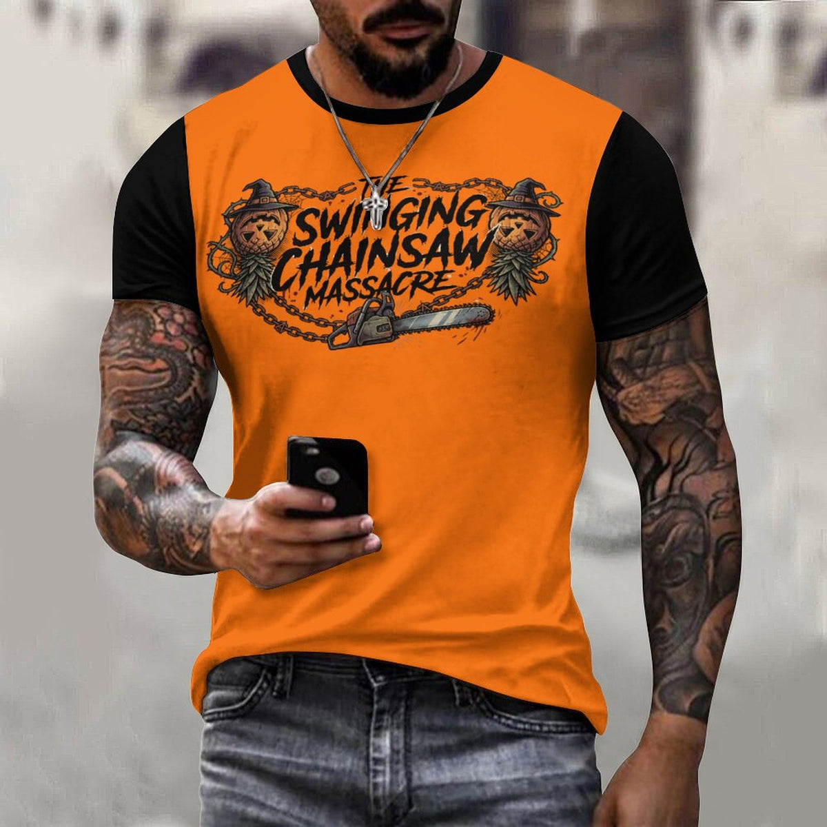 A man wearing a bright orange and black raglan t-shirt with a graphic print that reads 'The Swinging Chainsaw Massacre' featuring cartoon chainsaw and bear illustrations. He is holding a black smartphone and wearing gray jeans, set against a blurred gray background.