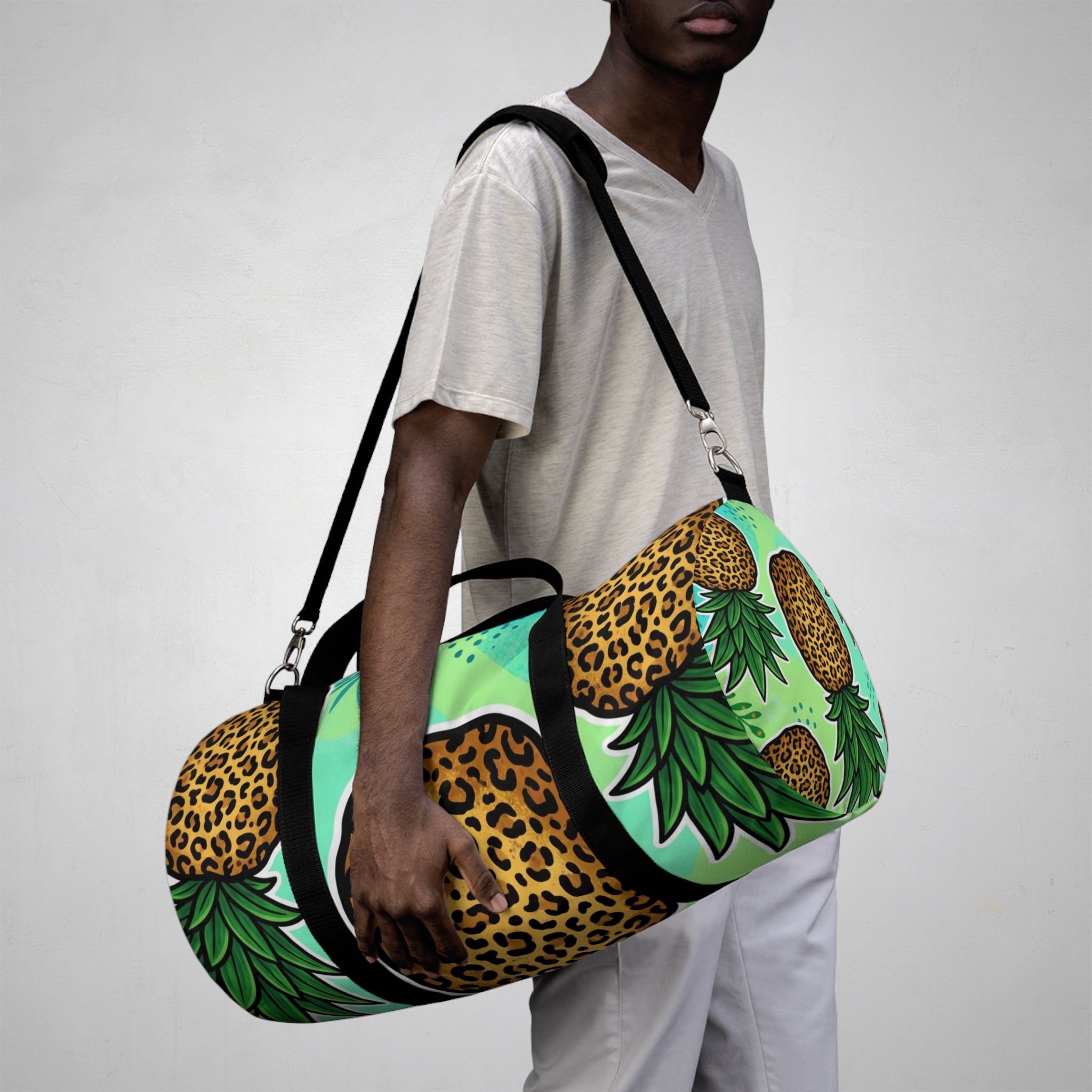 A person wearing a white t-shirt and white pants carries a green duffel bag decorated with a pineapple and leopard print pattern, set against a plain light gray background.