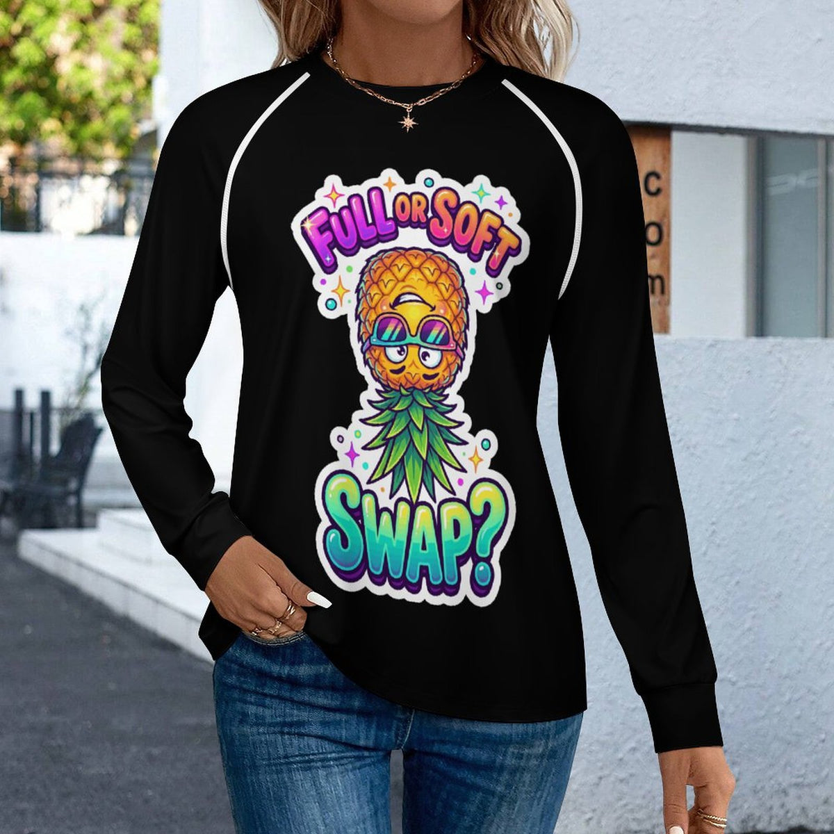 A woman wearing a black long-sleeve raglan shirt with a colorful pineapple graphic and the text 'FULL OR SOFT SWAP?' on the front, paired with blue jeans in an outdoor setting.