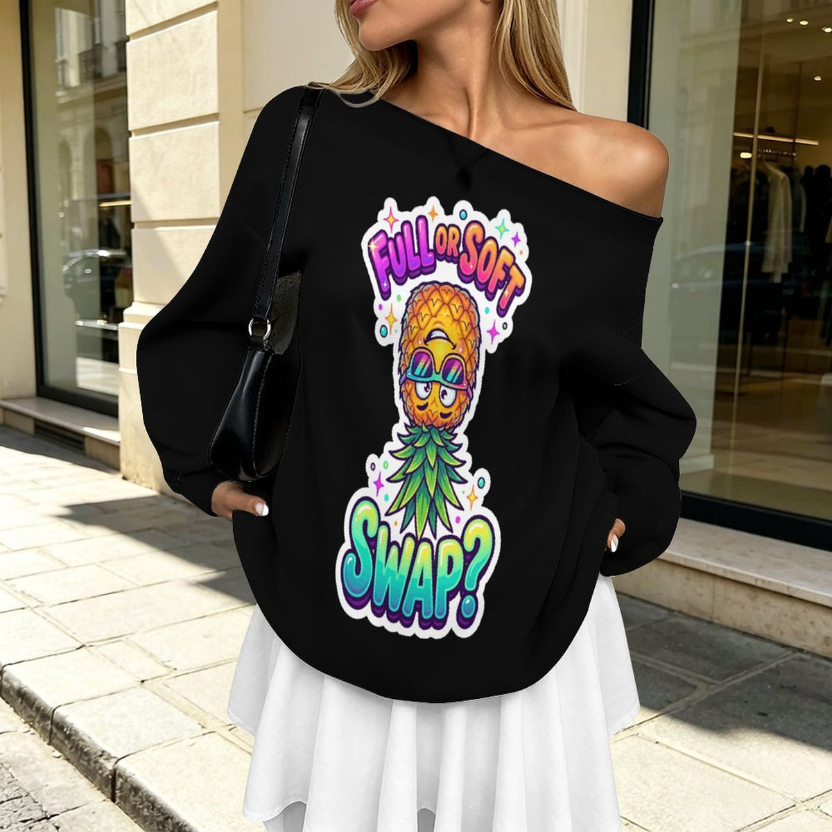 A woman wearing a black off-the-shoulder sweatshirt with a colorful pineapple graphic and the text 'FULL OR SOFT SWAP?' in a real outdoor storefront setting.