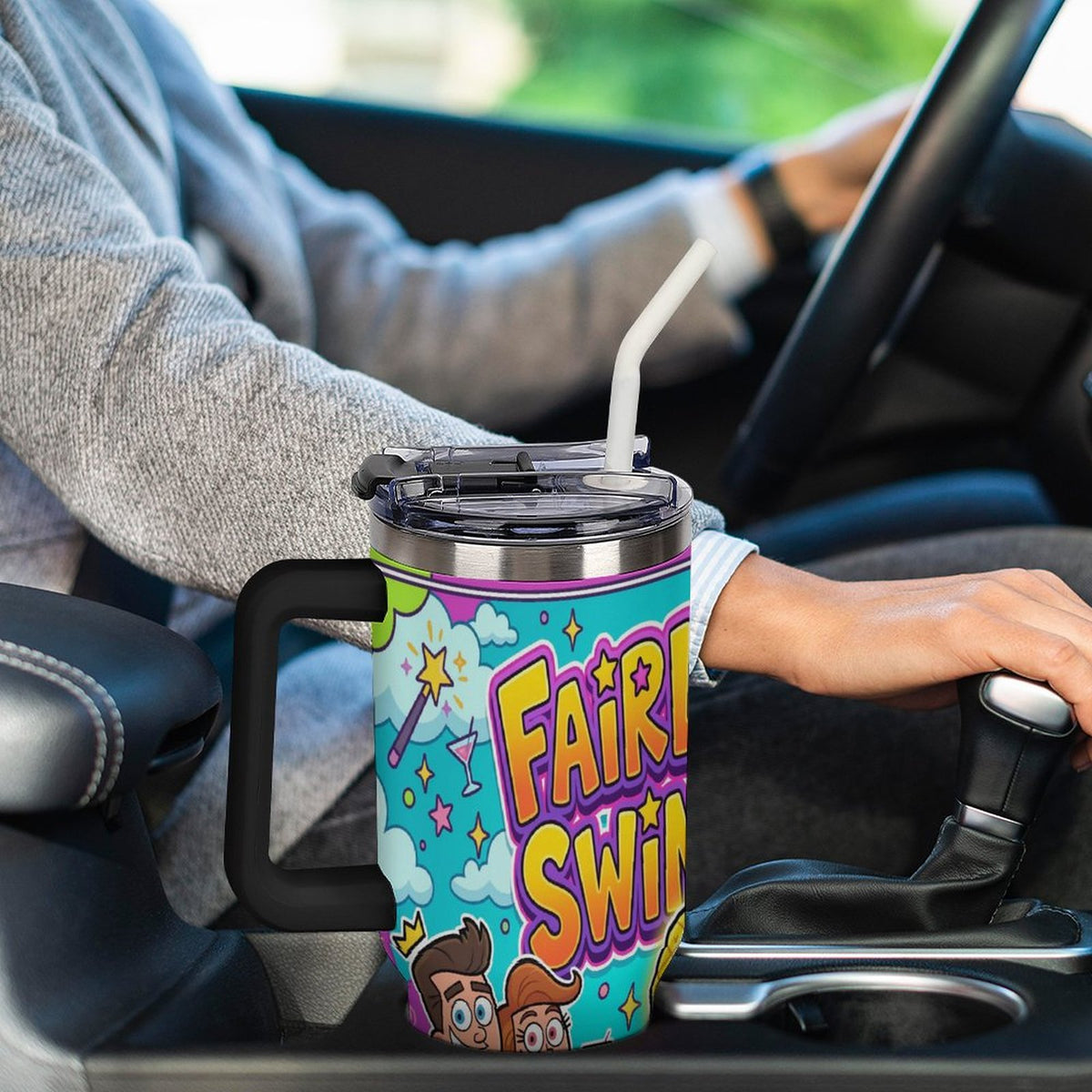 A person driving a car holds a handle on a large insulated tumbler cup. The cup has a colorful cartoon-style graphic with the text 'FAIRY SWIN' and a turquoise background.