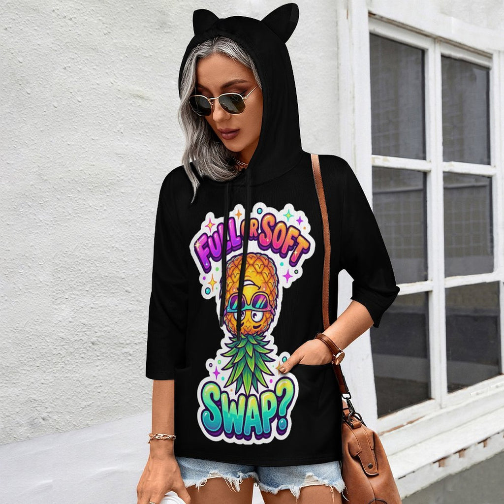 A woman wearing a black hoodie with cat ears and a colorful pineapple graphic that reads 'FULL SOFT SWAP?' stands outdoors against a white wall and window.
