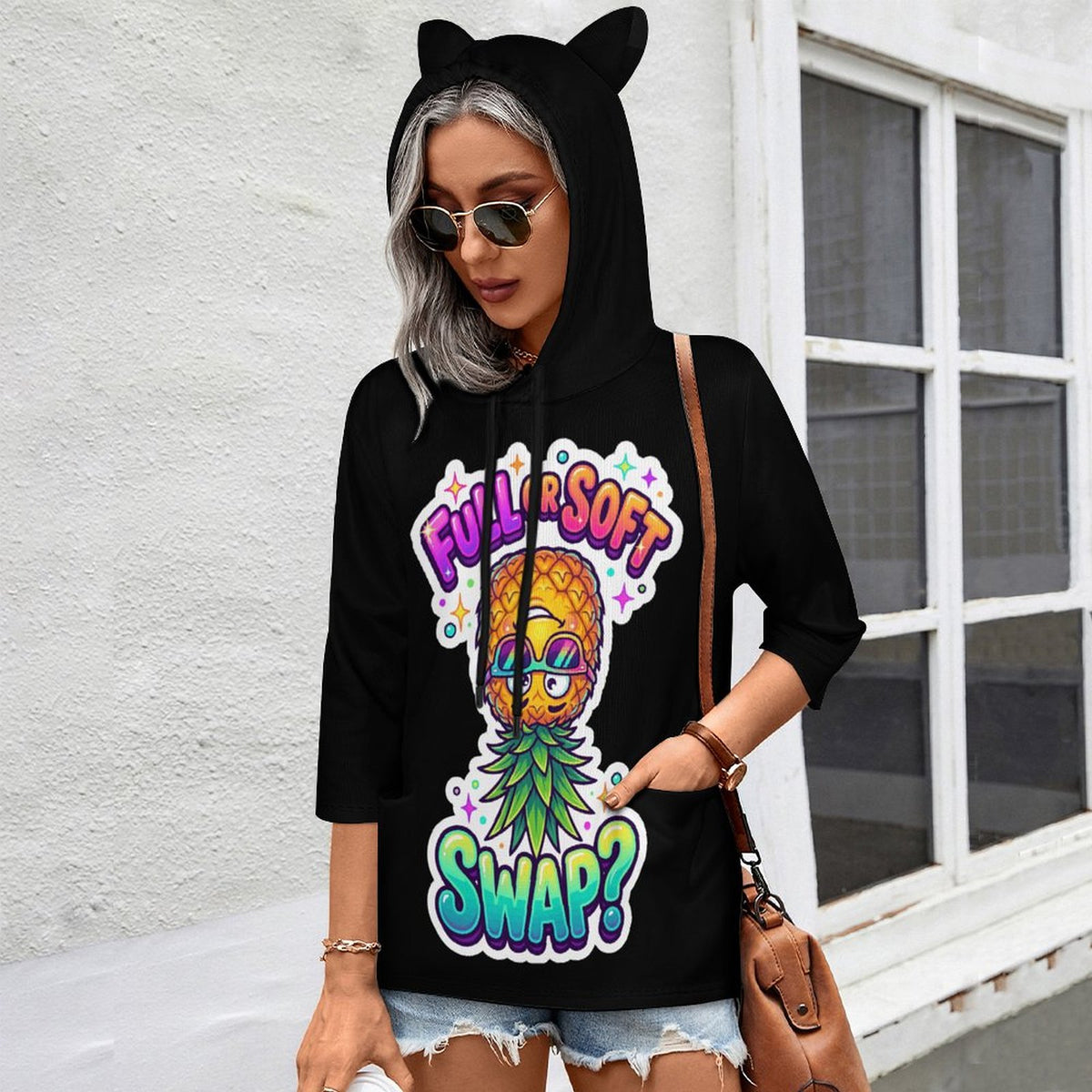 A woman wearing a black hoodie with cat ears and a colorful pineapple graphic that reads 'FULL SOFT SWAP?' stands outdoors against a white wall and window.