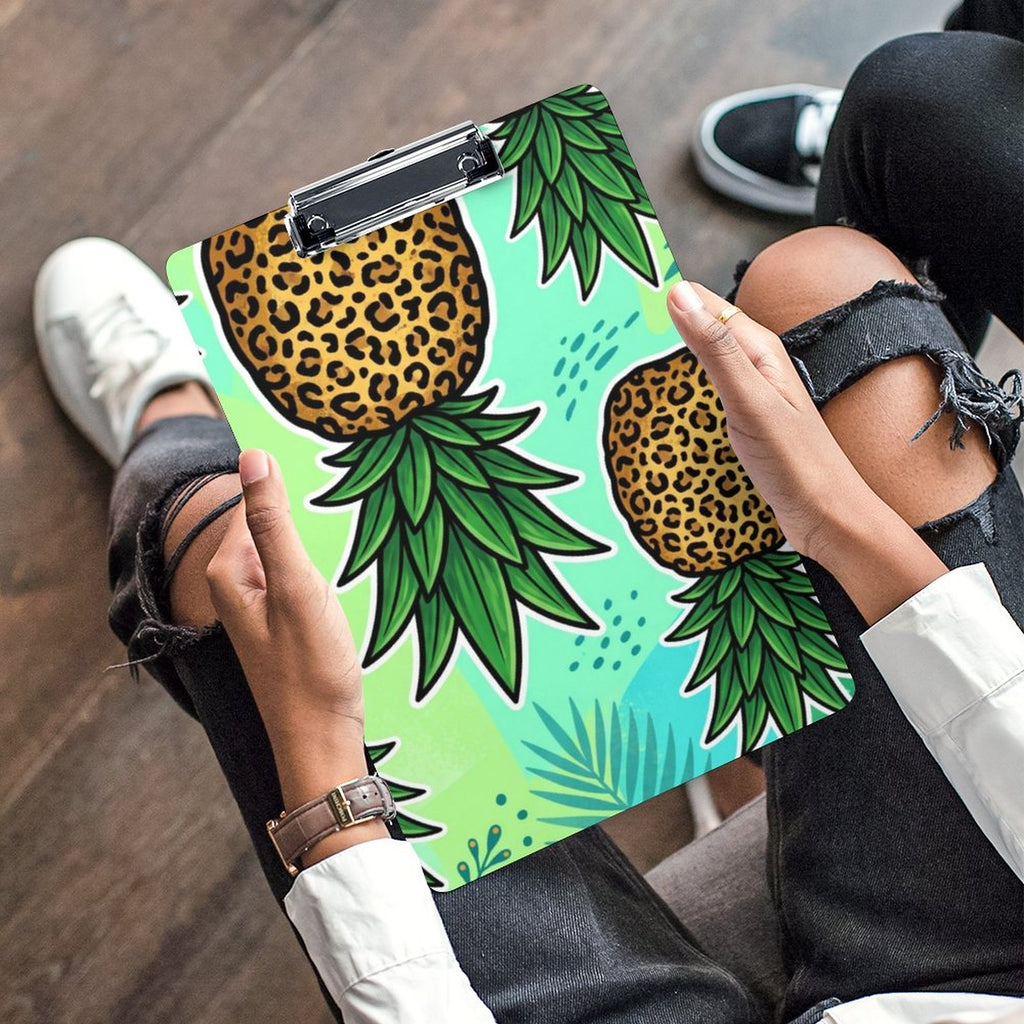 A person holding a clipboard with a colorful pineapple print, wearing ripped black jeans, white sneakers, and a white long-sleeve shirt, sitting on a wooden floor.