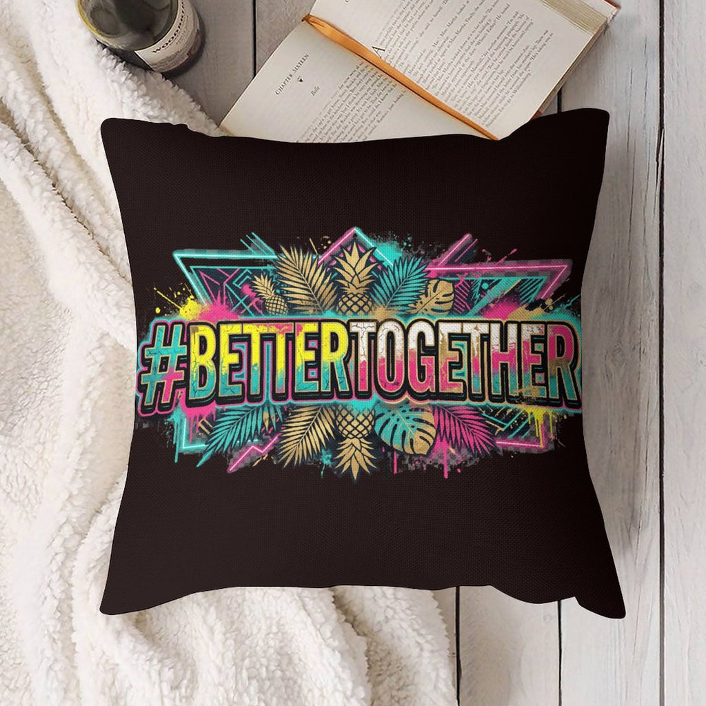 A square black throw pillow with a colorful graphic design featuring the text '#BETTERTOGETHER' in neon pink, blue, and yellow, surrounded by tropical leaves. The pillow is styled in a cozy flat-lay setting on a white textured blanket with an open book and a pen in the background.