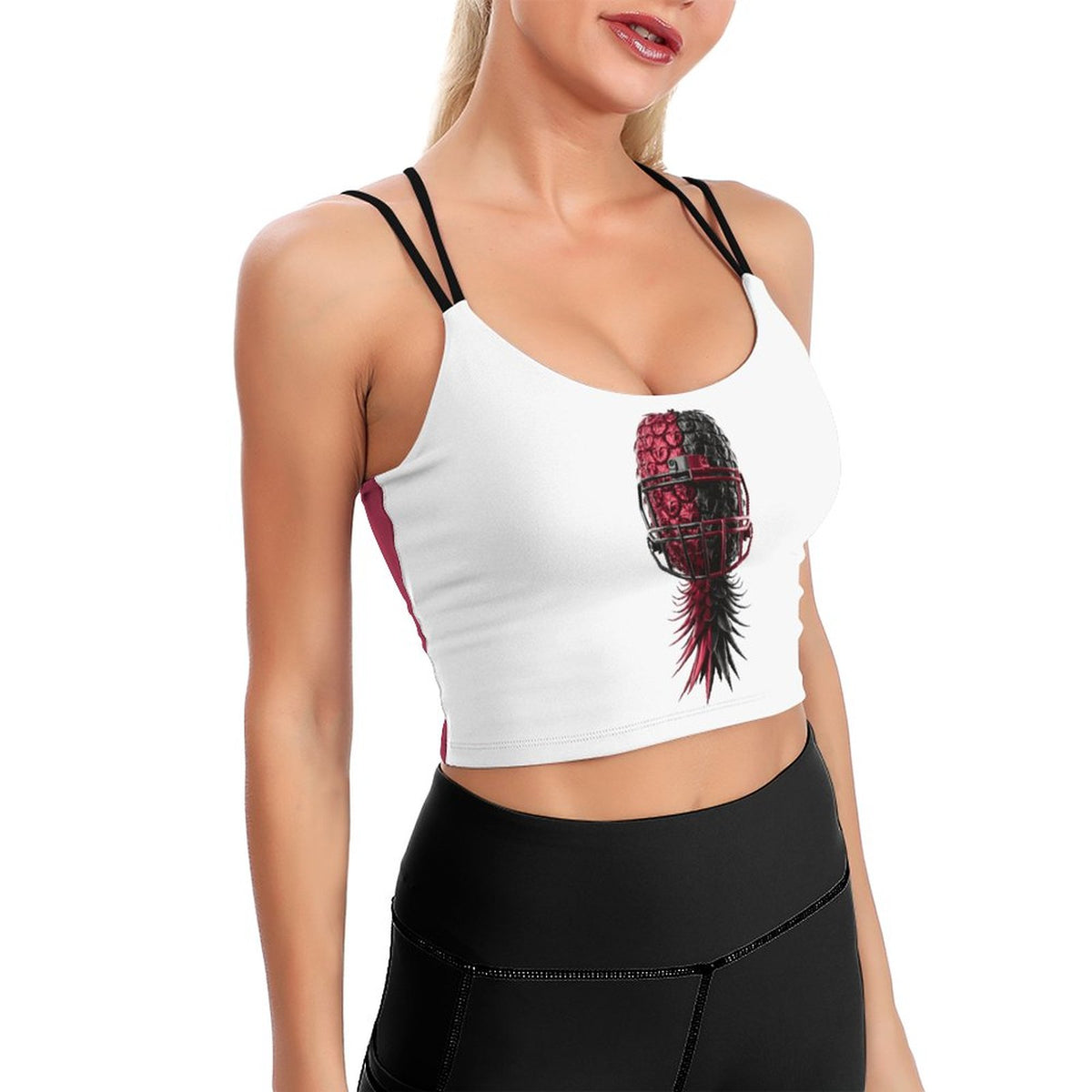 A woman wearing a white athletic crop top with black double-strap details and a graphic of a pineapple styled like a football helmet in red and black, paired with black leggings, against a plain white background.