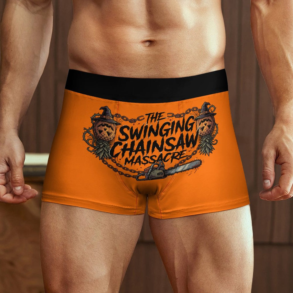 A close-up of a male model wearing bright orange boxer briefs with a black waistband and a large graphic print reading 'THE SWINGING CHAINSAW MASSACRE' with chainsaw and ape illustrations.