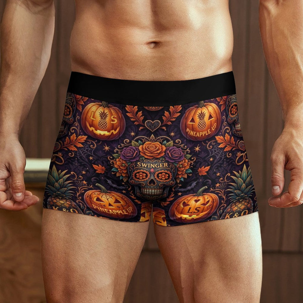 A close-up of a man's lower torso wearing boxer briefs with a colorful Day of the Dead style Halloween print featuring jack-o'-lanterns, roses, a sugar skull, and the words 'Swinger' and 'Pineapple'.