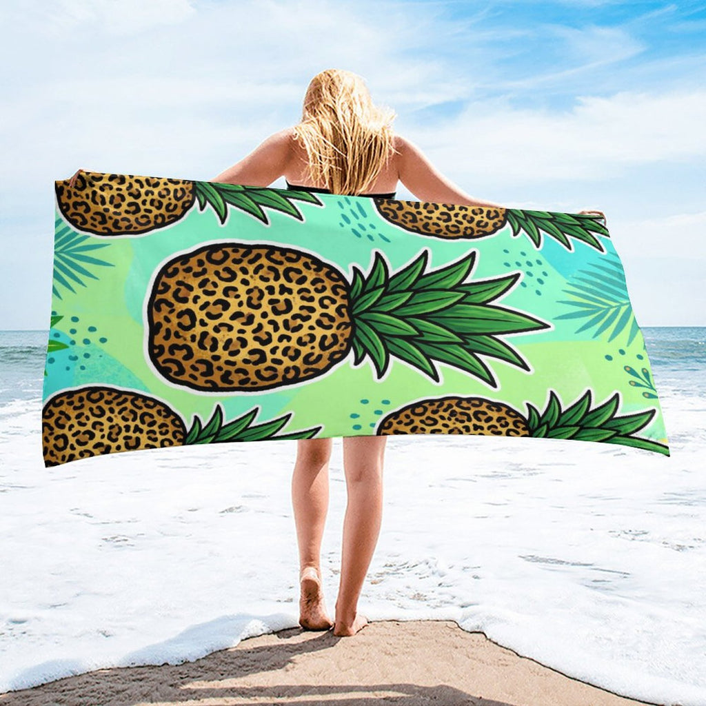 A woman stands on a sandy beach holding up a large beach towel. The towel features a vibrant tropical design with green palm leaves and bold leopard-print pineapples on a light mint green background.