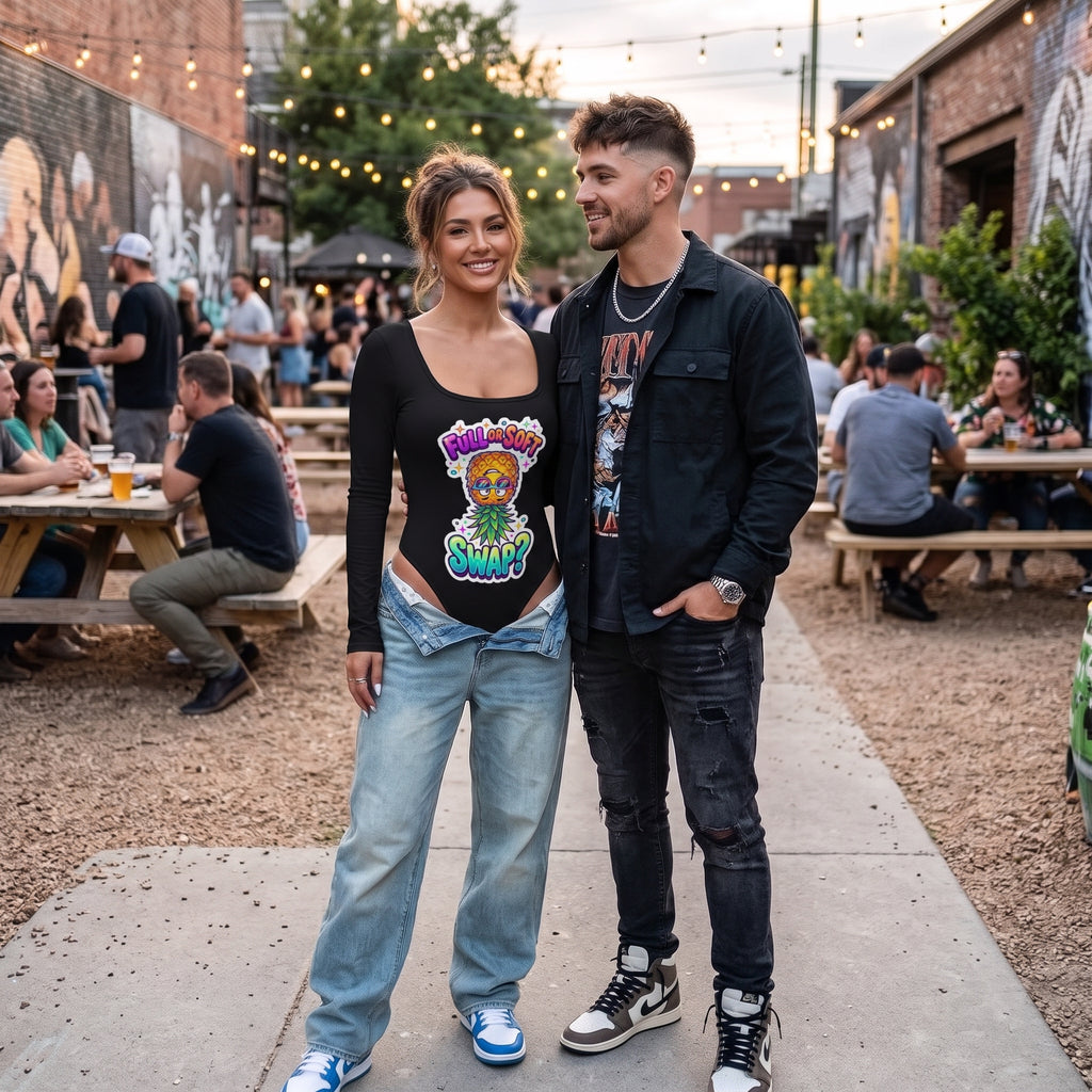 A couple standing outdoors at a casual brewery or patio setting, with the woman wearing a black scoop-neck graphic t-shirt featuring a colorful Hawaiian-style design and light-wash jeans, and the man wearing a black jacket over a graphic tee and dark jeans.